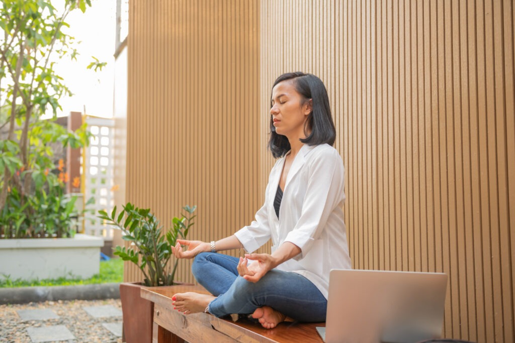 Woman sitting cross-legged meditating outdoors near wooden wall and plants